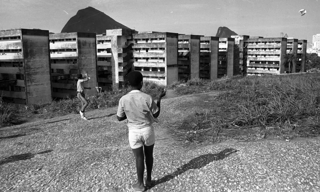 Dois meninos brincando em um terreno aberto com prédios ao fundo.