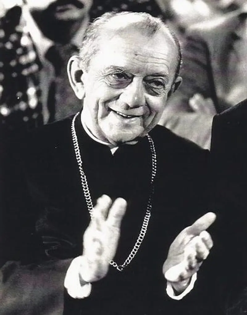 Homem sorridente em trajes religiosos levanta dedos em gesto expressivo, multidão ao fundo.