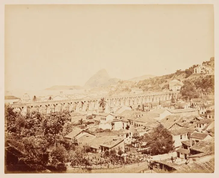 Cidade antiga com ponte aqueduto central, edifícios coloridos à esquerda e montanha verde ao fundo.