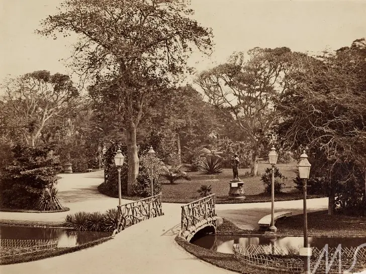 Jardim antigo com árvores densas, caminho curvado e monumento erguido.