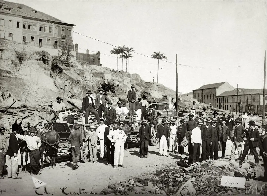Cena urbana histórica com pessoas caminhando e carruagens paradas.