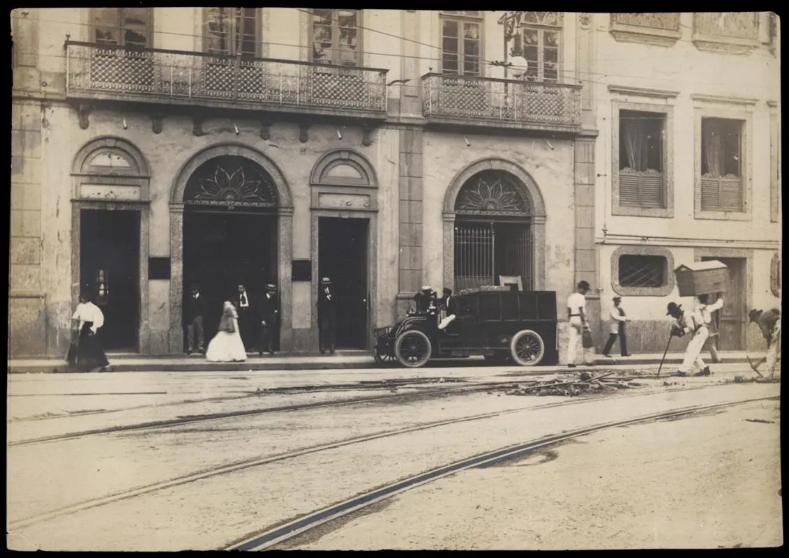 Edifício clássico com pessoas em trajes formais e carro antigo.