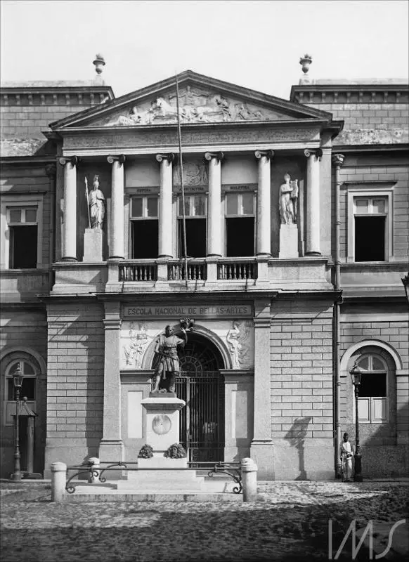Edifício histórico com estátuas femininas segurando espadas e instrumentos musicais, balcão com janelas e placa da Escola Nacional de Belas Artes.