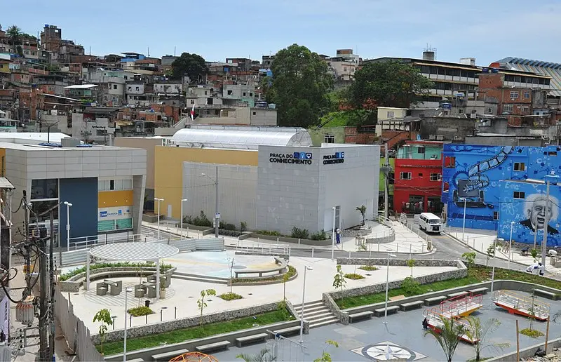 Uma praça urbana com edifícios modernos e favela colorida ao fundo.