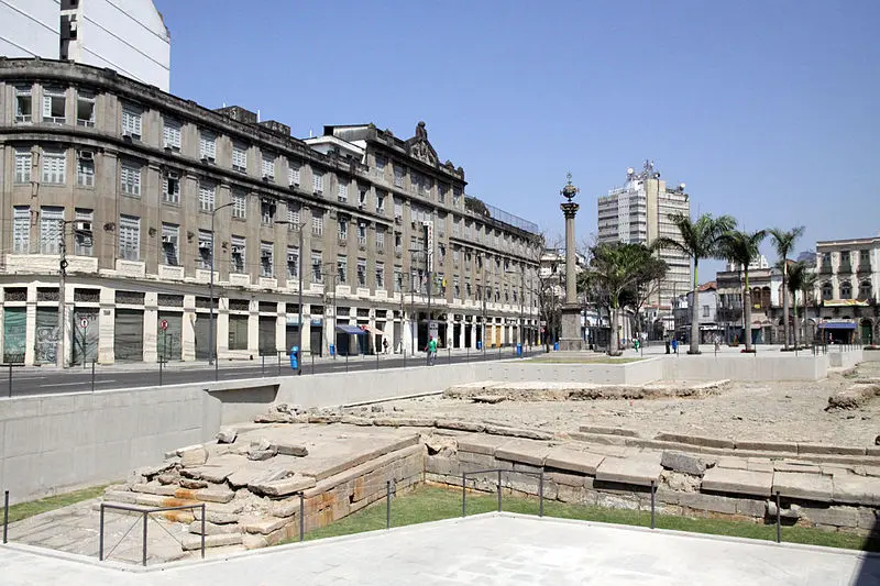 Imagem urbana com edifícios, pedra e concreto, palmeiras e céu azul claro.