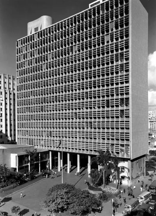 Edifício modernista com torre e cúpula, árvore e pessoas caminhando.