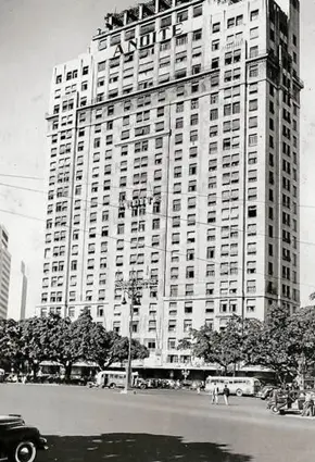 Edifício moderno em cruzamento urbano, árvores e pessoas caminhando.