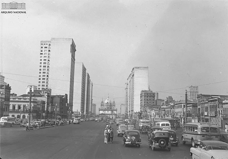 Avenida movimentada com edifícios modernos e antigos, igreja com cúpula, carros antigos e céu nublado.