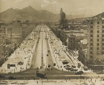 Avenida movimentada com construções antigas e modernas, veículos circulando e montanha ao fundo.