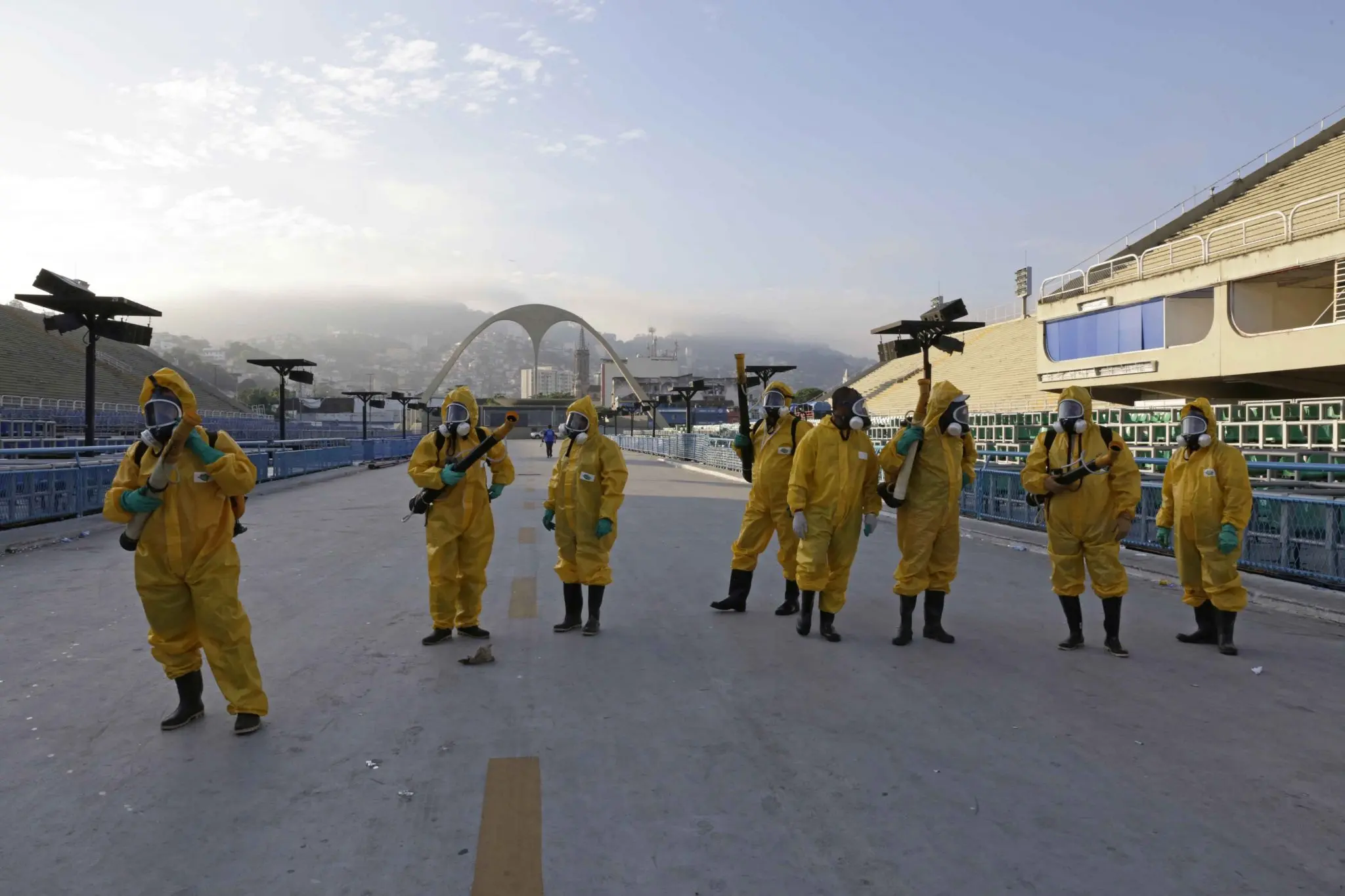 Pessoas em trajes de proteção em sambódromo, arquibancada e ponte ao fundo.