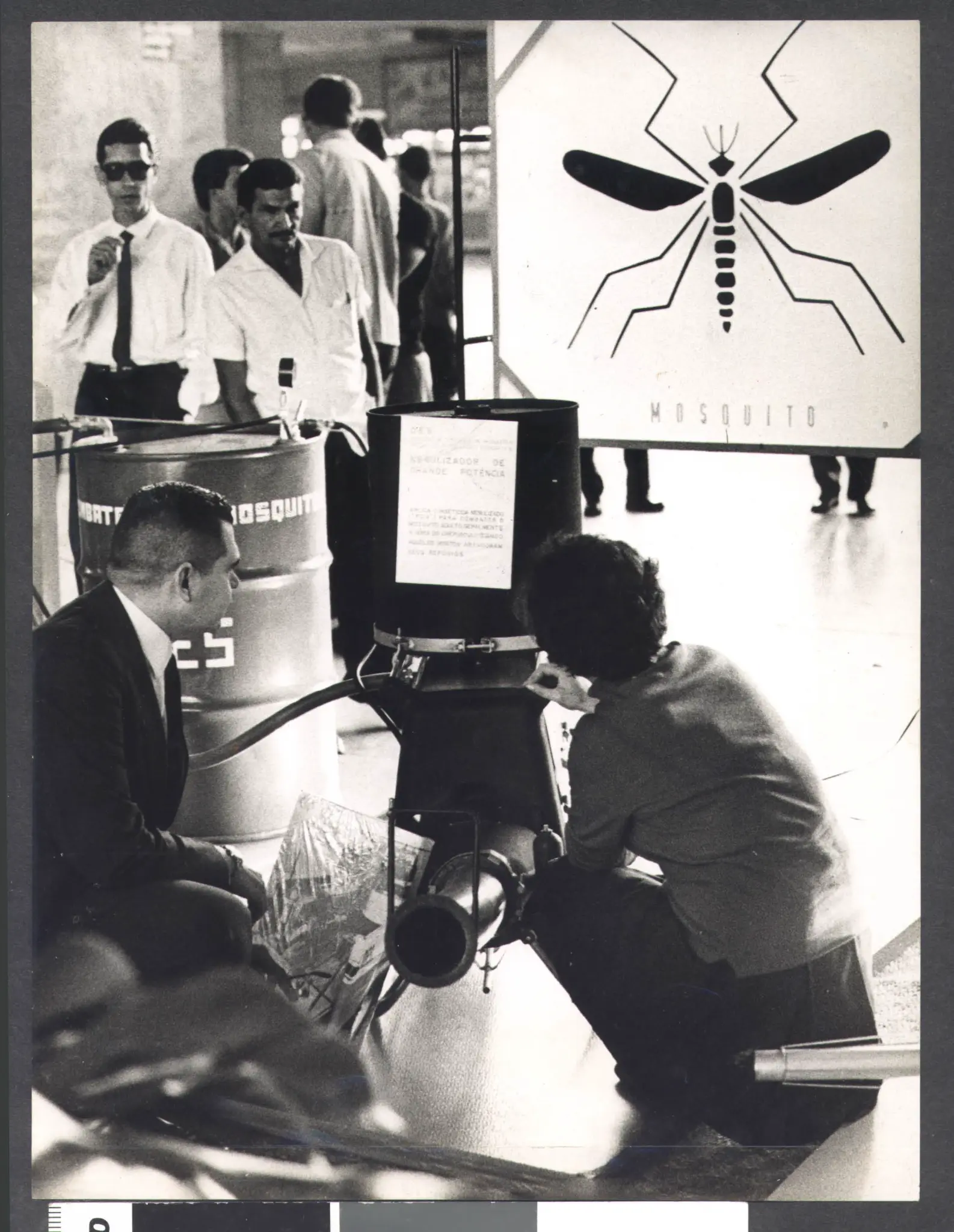 Pessoas em trajes formais observam inseto; dois ajoelhados examinam dispositivo com "MOSQUITO" rotulado.
