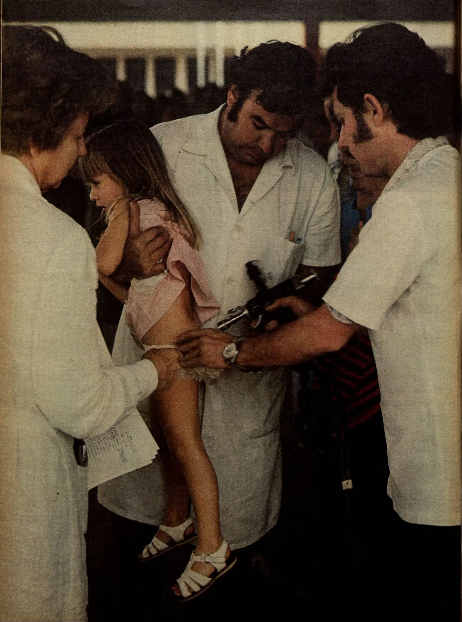 Uma cena médica com mulher segurando criança, homem administrando injeção e outros observando.