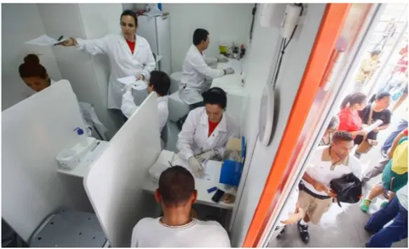 Ambiente médico com profissionais distribuindo documentos e pacientes aguardando atendimento.