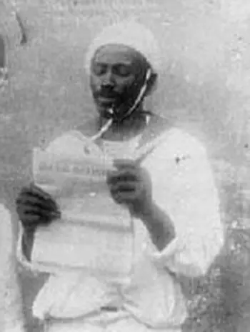 Homem em trajes tradicionais segurando documento, fundo desfocado.