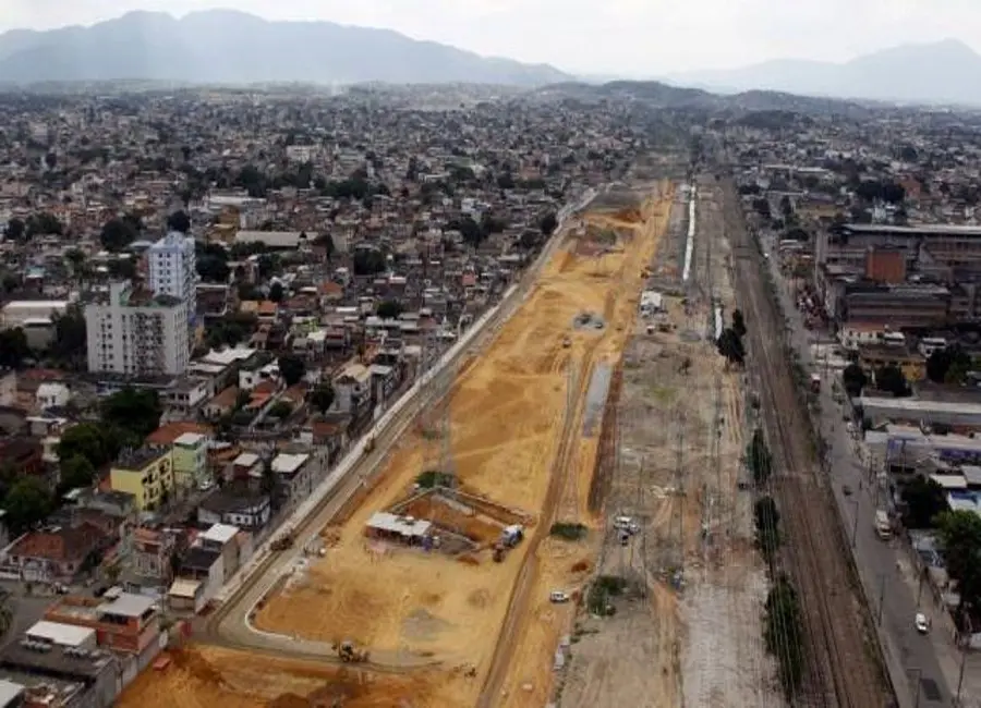 Área urbana em construção com edifícios residenciais, espaço aberto, via férrea e montanhas ao fundo.