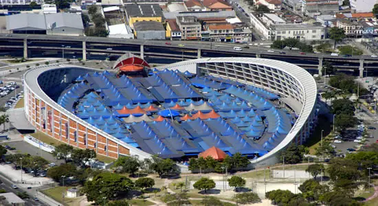 Estádio urbano com arquibancadas azuis e vermelhas, telhados azuis e edifícios ao fundo.