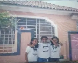 Três pessoas sorridentes em frente a prédio residencial amarelo.