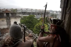 Dois indivíduos em urbano, um com chapéu de malha e outro em trajes tradicionais indígenas segurando arco e flecha, em construção abandonada com telhados danificados e edifício moderno ao fundo.