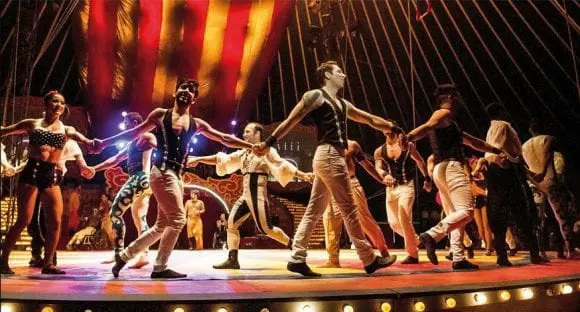 Dançarinos em palco com iluminação dramática, tons de vermelho e dourado.