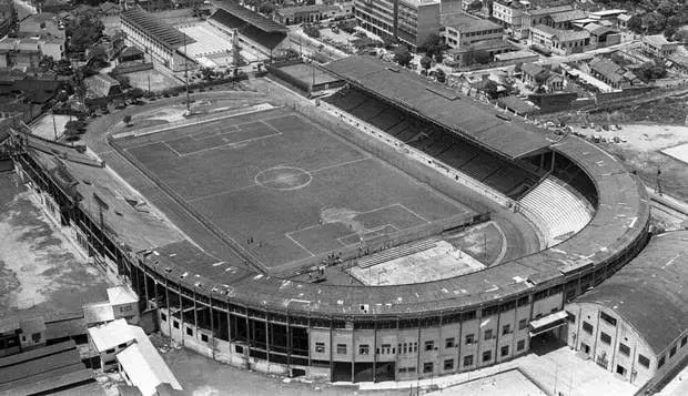 Estádio em preto e branco com arquibancadas e gramado verde.