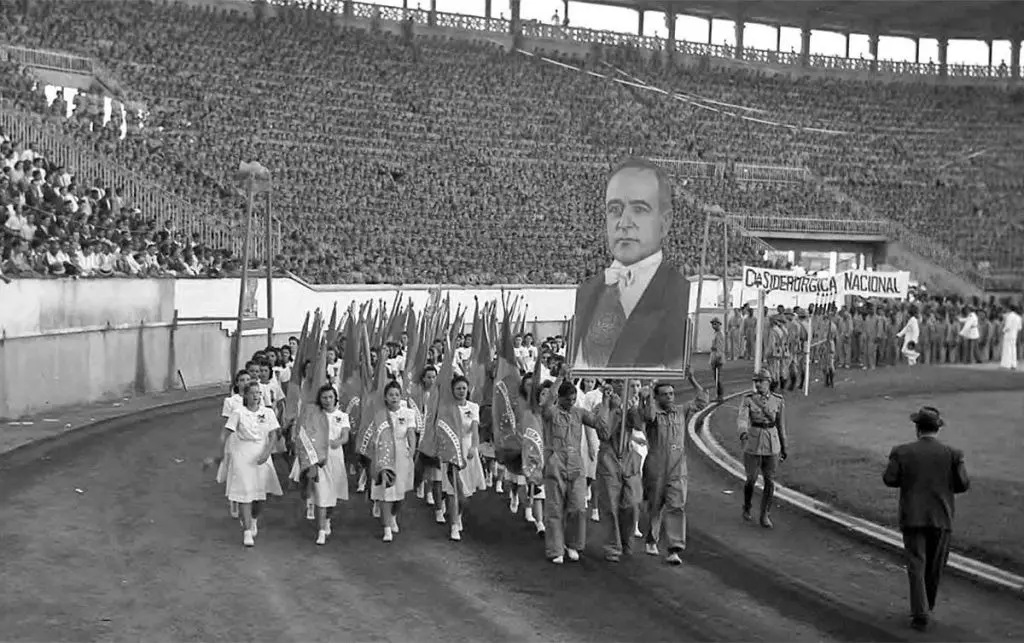 Desfile em estádio com figura humana destacada e procissão de bandeiras.