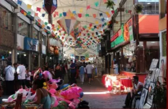 Mercado tradicional com pessoas caminhando e comprando, barracas coloridas e decoração festiva.
