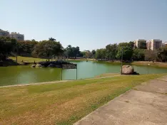 Parque com lago, gramado verde, banco e prédio moderno.