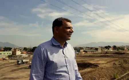 Homem sorridente em camisa xadrez azul, em local com vegetação e construção, com casas e montanha ao fundo.