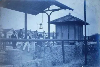 Estação ferroviária antiga em preto e branco, com plataforma elevada e pessoas esperando.