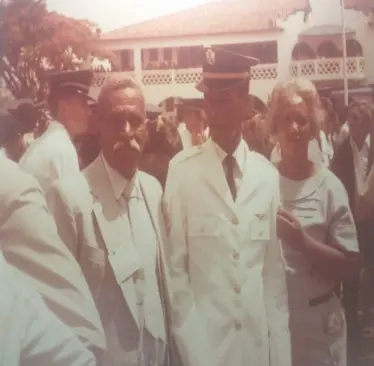 Grupo de pessoas em evento público, com homem em terno e distintivo, mulher em vestido claro, edifício ao fundo.