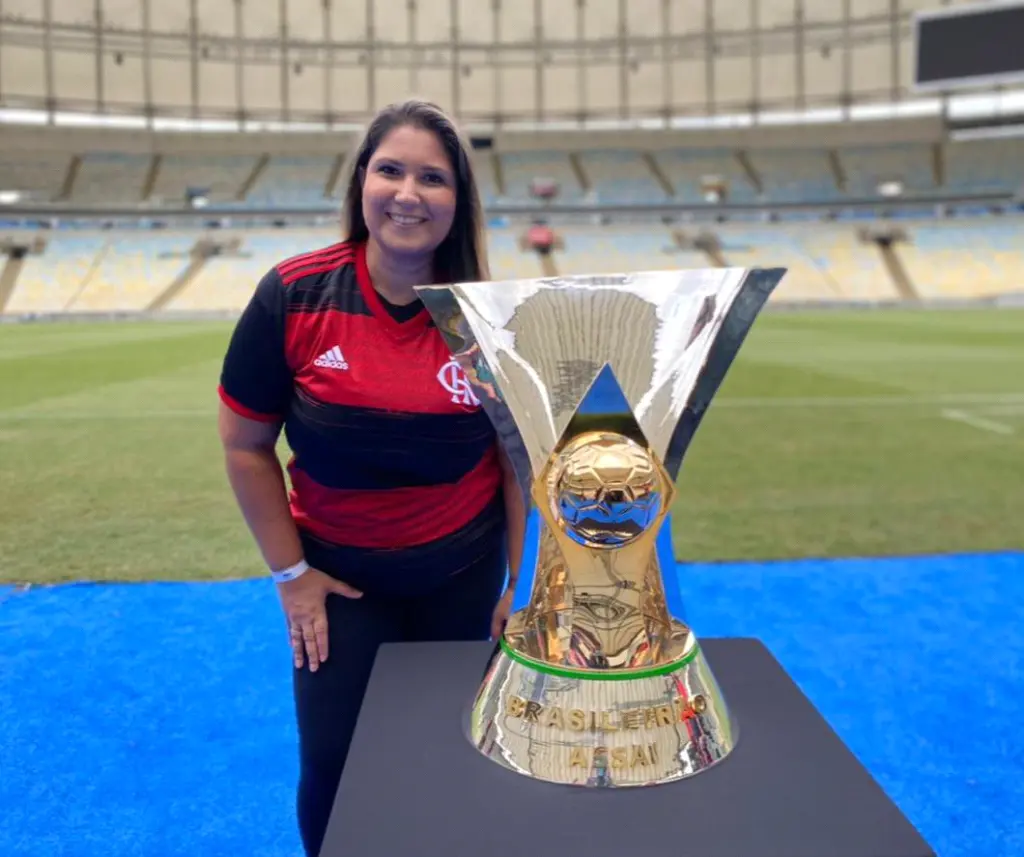Mulher sorridente com camisa do Flamengo em estádio vazio, segurando troféu dourado.