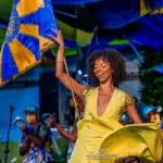 Mulher dançando samba com traje amarelo vibrante, segurando bandeira azul e amarela.