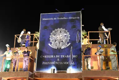 Desfile de escola de samba com cartaz sobre trabalho e previdência social.
