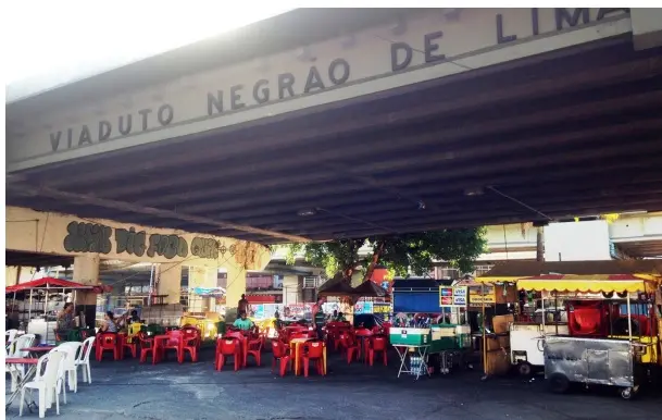 Mercado local sob viaduto com mesas vermelhas e barracas coloridas.