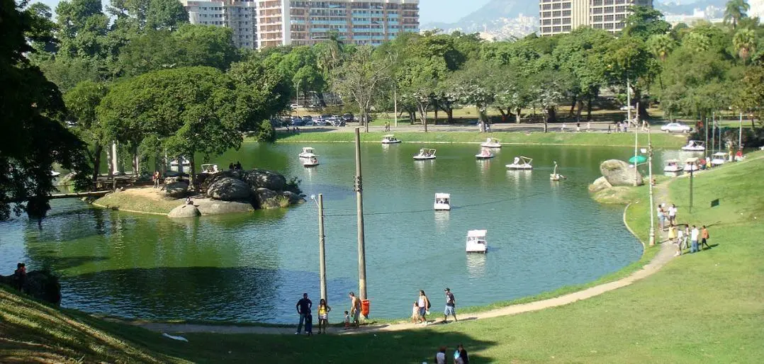 Parque com lago, árvores, rochas, edifícios altos, caminhantes e barcos.