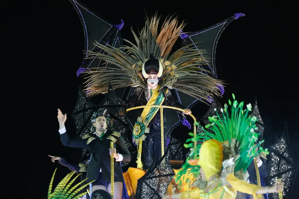 Figura em traje de carnaval com faixa presidencial e coroa de penas, segurando arco e flecha.