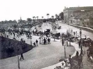 Praça histórica com pessoas caminhando e carruagens, gramado verde e construções de tijolos vermelhos.