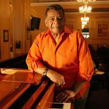 Homem sorridente em frente ao piano, sala com lustre pendente.