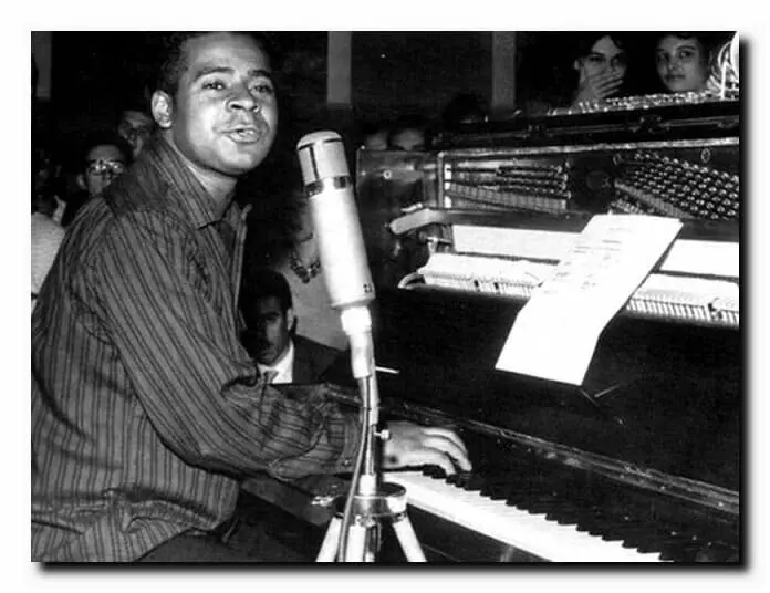 Homem cantando em estúdio perto de piano com partituras.