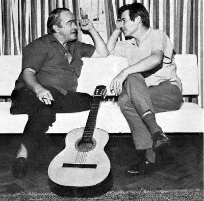 Dois homens sorridentes em conversa, guitarra clássica ao lado.