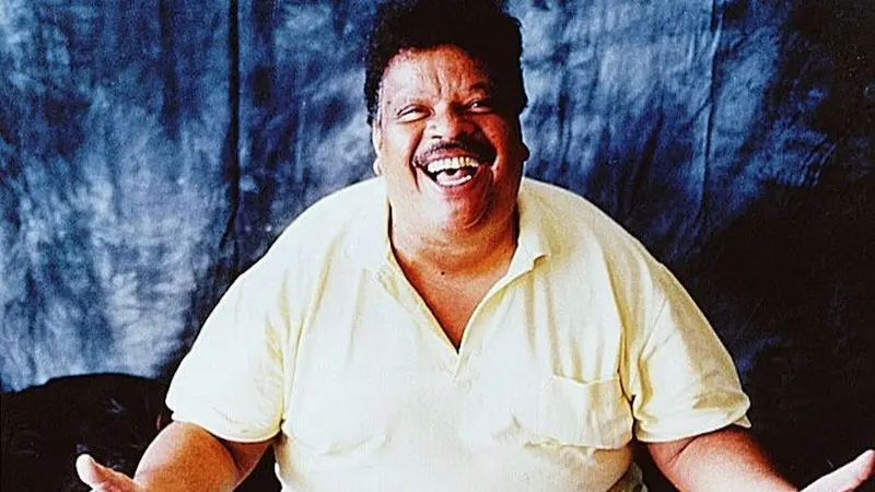 Homem sorrindo em fundo azul escuro, camisa amarela.