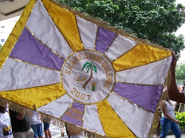 Bandeira colorida com design radial, palmeira e concha no centro, texto "GRANDE SAMAR COLOMBO" em 2007.