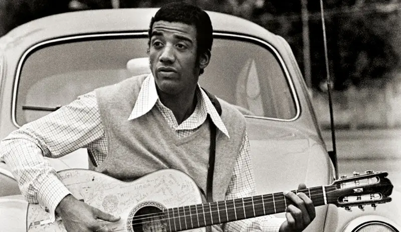 Homem com guitarra em frente a carro clássico, jardim ao fundo.