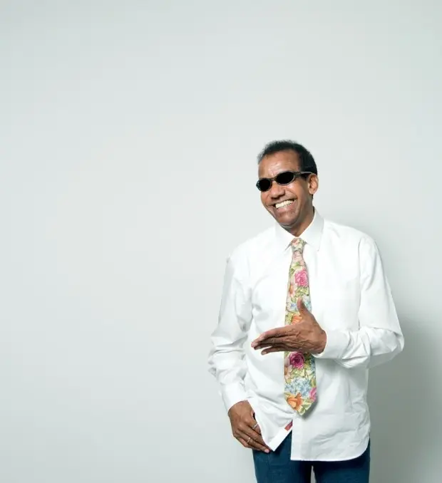 Homem sorridente em camisa branca com laço floral verde, rosa e amarelo, usando óculos escuros, fundo branco.
