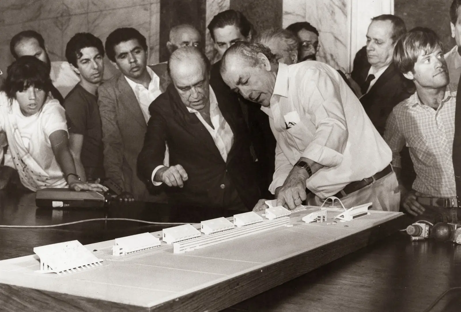 Pessoas reunidas em torno de uma maquete de edifícios, discutindo projeto arquitetônico.