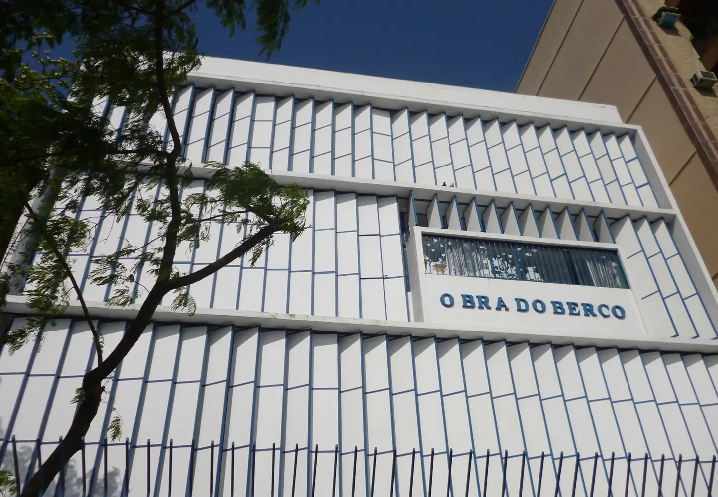 Edifício moderno com fachada branca e azul, árvore verde à esquerda, varanda metálica à direita, placa "O BRA DO BERCO" no centro.