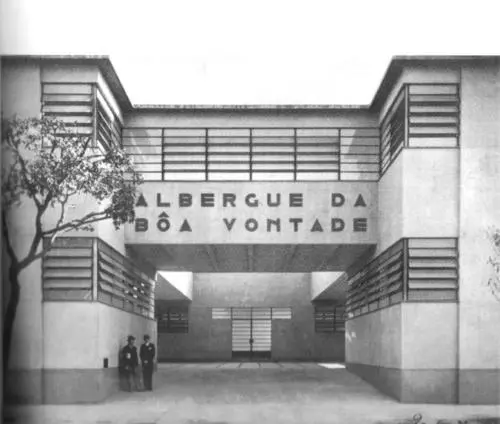 Edifício modernista com árvore, entrada arqueada e estrutura simétrica.