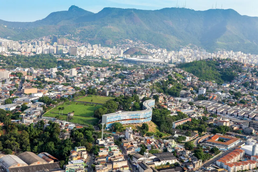 Cidade com vista panorâmica, áreas verdes, edifícios, estradas, montanha verde, estádio distante, sol brilhante, céu azul claro.