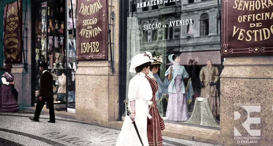 Rua animada do início do século XX, com loja de roupas femininas, duas mulheres elegantes caminhando e um homem se aproximando.