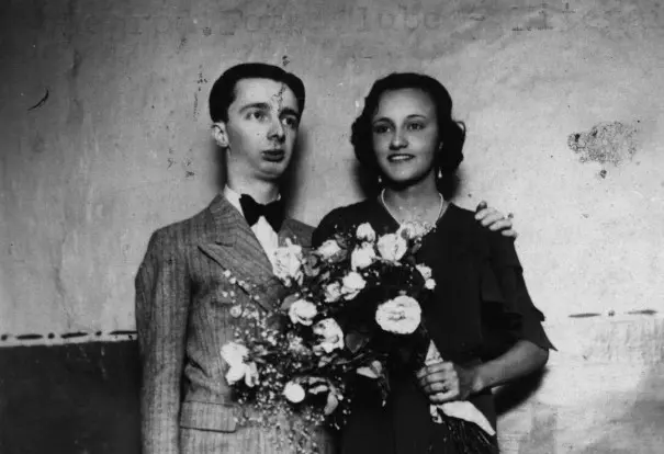 Casal sorridente em foto preto e branco, mulher com buquê e vestido escuro, homem em terno clássico.